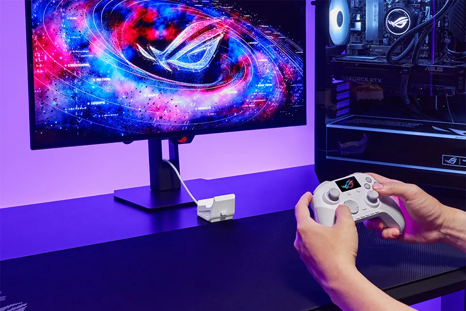 De witte ROG Raikiri II Pro PC in twee handen, met de oplaadstandaard en dongle naast de monitor